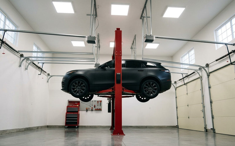 Black SUV raised high on a red two-post lift in a tall garage with high ceilings